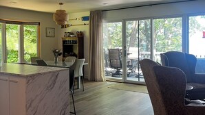 A spacious living area with a marble kitchen island, a dining table with chairs, a large window with a view of greenery, and a patterned armchair.