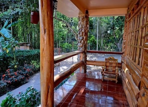 Un balcón de madera con un banco, rodeado de exuberante vegetación y un piso de baldosas.