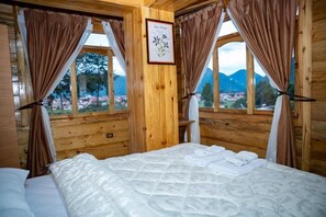 A wooden cabin room with a bed, curtains, a framed botanical print, and a window view of a forest and mountains.