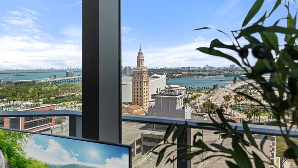 A view from a window overlooking a cityscape with a prominent building, a bridge, and a body of water. - 501 First 2001 Ocean View Corner Two Bedroom (Miami)