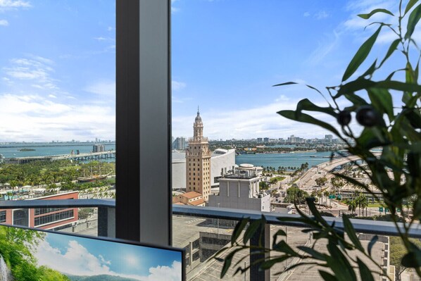A view from a window overlooking a cityscape with a prominent building, a bridge, and a body of water. - 501 First 2001 Ocean View Corner Two Bedroom (Miami)