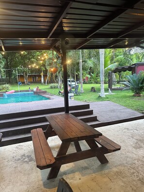 A covered outdoor area with a pool, picnic table, and steps leading to a grassy area with trees and string lights.