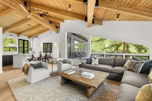 A spacious living area with a wooden ceiling, large windows, a sofa, a coffee table, and a kitchen in the background.