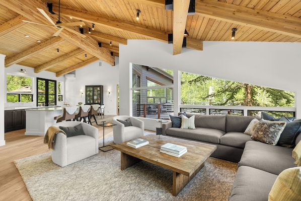 A spacious living area with a wooden ceiling, large windows, a sofa, a coffee table, and a kitchen in the background.