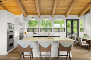 A modern kitchen with white cabinets, stainless steel appliances, a wooden ceiling, and large windows.