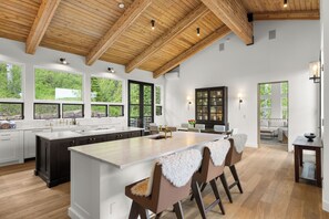 A spacious kitchen with a large island, wooden ceiling beams, and large windows offering views of greenery. - Elegant Snowmass Village Family Escape W/ Hot Tub, 2 Decks, Luxury Finishes | Snowmass Classic (Snowmass Village)