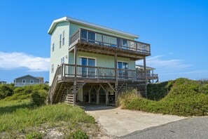 A two-story beachfront house with a balcony, a staircase leading to the entrance, and a sandy driveway. - South Nags Head 426: Semi-Oceanfront, Ocean Views, Hot Tub (Nags Head)