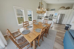 A dining area with a wooden table and chairs, a chandelier, and a kitchen in the background. - South Nags Head 426: Semi-Oceanfront, Ocean Views, Hot Tub (Nags Head)