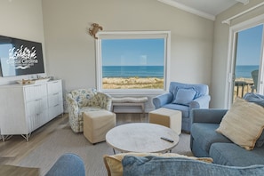 A living room with a large window overlooking a beach, a white cabinet, and a blue sofa. - South Nags Head 426: Semi-Oceanfront, Ocean Views, Hot Tub (Nags Head)