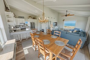 A spacious living area with a dining table, chairs, a blue sofa, and a view of the beach. - South Nags Head 426: Semi-Oceanfront, Ocean Views, Hot Tub (Nags Head)