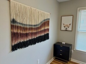Une pièce avec une tapisserie accrochée au mur, un tableau encadré, une commode de couleur sombre et une fenêtre avec des stores.