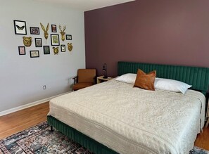 Une chambre à coucher avec un grand lit, une tête de lit verte, une table de chevet, une chaise et des œuvres d’art murales.
