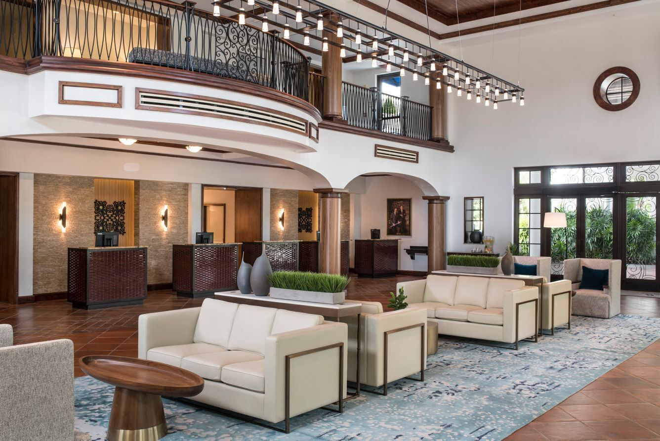 A spacious lobby with a curved reception desk, multiple seating areas, and a balcony with a railing.