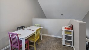 Huone, jossa on valkoinen pöytä, värikkäät tuolit, kirjahylly ja kalteva katto.