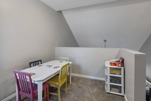 Ein Zimmer mit einem weißen Tisch, bunten Stühlen, einem Bücherregal und einer schrägen Decke.