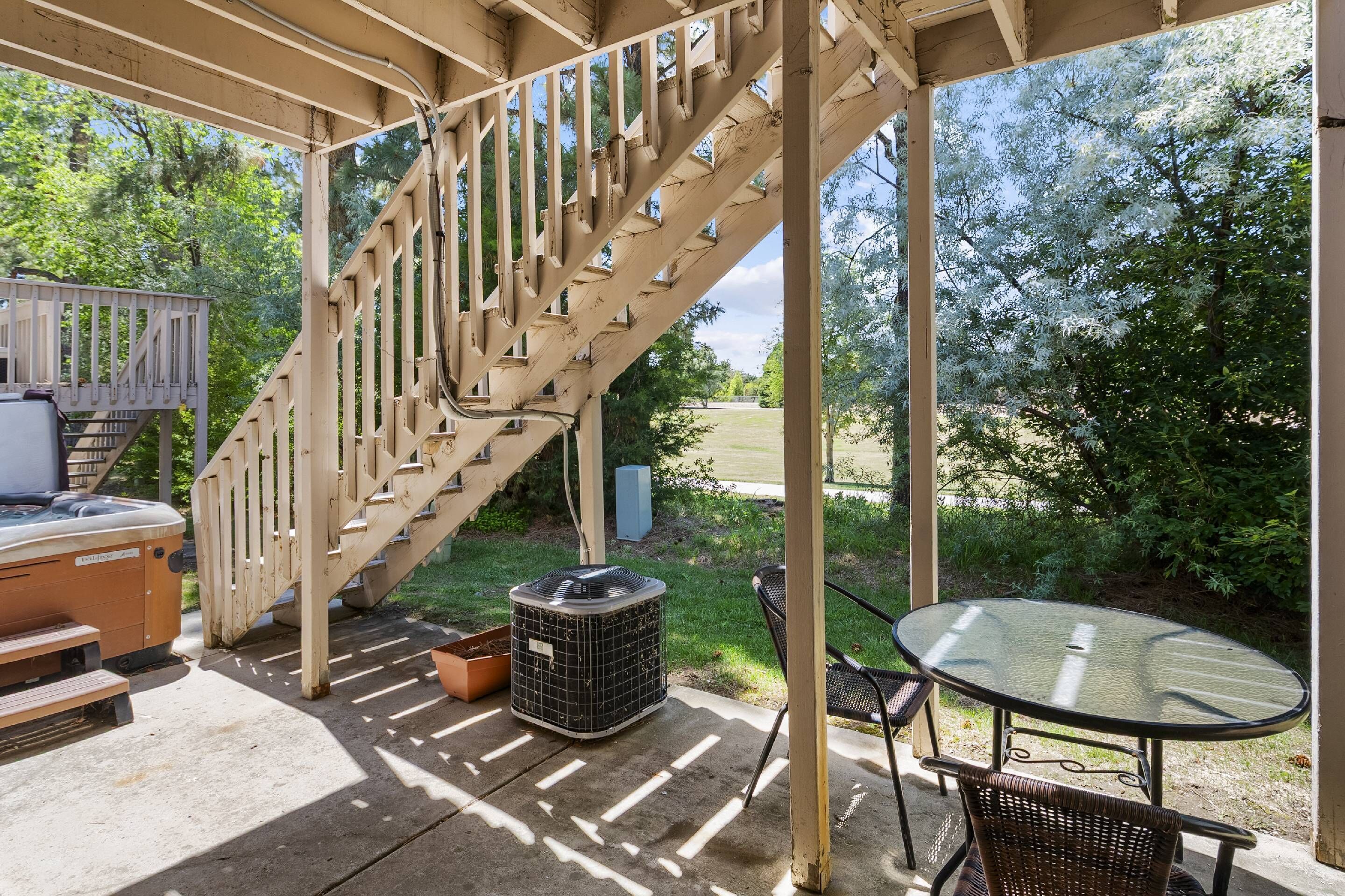 A covered outdoor area with a wooden staircase, a hot tub, a glass table, and chairs.
