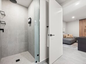 Ein modernes Badezimmer mit Duschkabine aus Glas, einer weißen Tür und einem grauen Sofa im angrenzenden Wohnbereich.