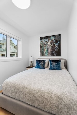 Ett sovrum med en stor säng, ett fönster med utsikt över hus och en tavla på väggen.