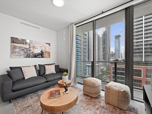 Ein modernes Wohnzimmer mit grauem Sofa, rundem Holztisch und einem großen Fenster mit Blick auf die Stadt.