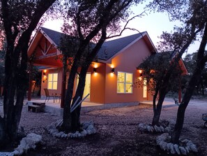 A house with a sloped roof, a porch with seating, and a well-lit interior. - Delightful 1-bedroom cottage with AC in Dripping Springs (Dripping Springs)
