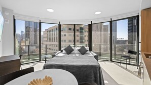 Ein modernes Schlafzimmer mit einem großen Bett, einem kleinen Tisch und einem Sessel. Es verfügt über einen Balkon mit Blick auf Stadtgebäude.