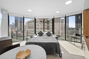 Ein modernes Schlafzimmer mit einem großen Bett, einem kleinen Tisch und einem Sessel. Es verfügt über einen Balkon mit Blick auf Stadtgebäude.