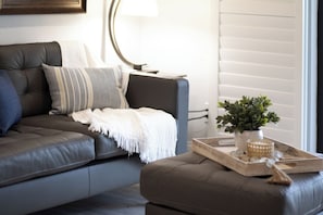 A modern living room with a grey sofa, a white throw blanket, a potted plant, and a tray with a broom. - Direct Oceanfront Condo | Balcony, Heated Pool + Putting Green 🍹🏝️ (Cocoa Beach)