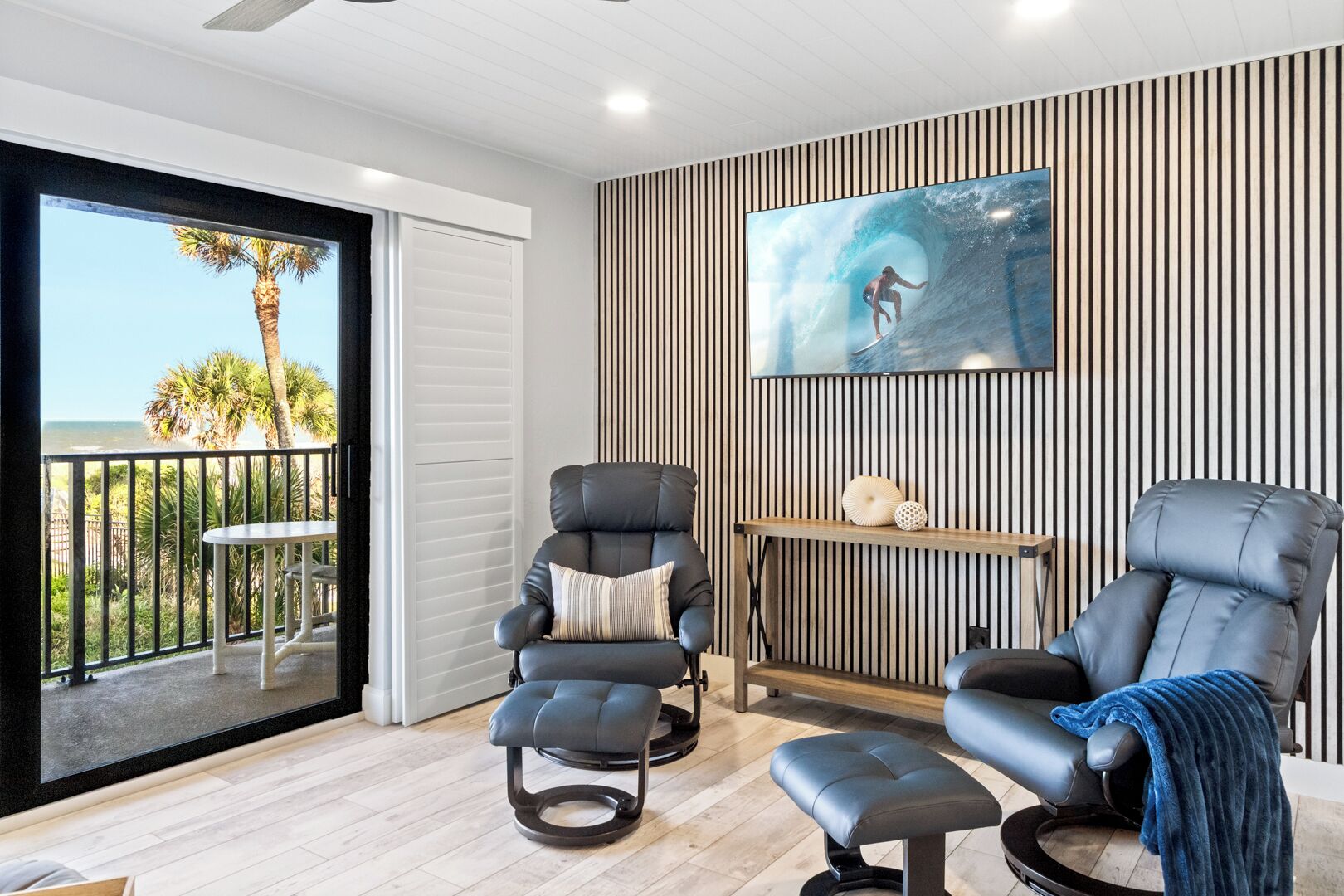 A modern living room with a large window, two reclining chairs, a small table, and a wall-mounted TV.