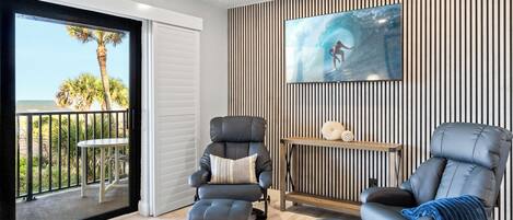 A modern living room with a large window, two reclining chairs, a small table, and a wall-mounted TV.