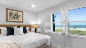 Ein Schlafzimmer mit einem großen Bett, einem Nachttisch, einer Lampe, einer Bank, einem Gemälde und einem Fenster mit Blick auf den Strand.
