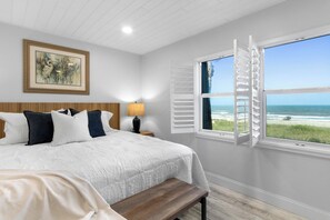 A bedroom with a large bed, a nightstand, a lamp, a bench, a painting, and a window with a view of the beach. - Direct Oceanfront Condo | Balcony, Heated Pool + Putting Green 🍹🏝️ (Cocoa Beach)