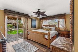 Ein Schlafzimmer mit Bett, Deckenventilator, Nachttisch, Korbsessel, Holzboden und Blick auf einen Garten durch eine Schiebetür.