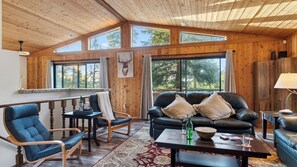 Ein geräumiger Raum mit Holzbalkendecke, großen Fenstern, einem schwarzen Ledersofa, einem Holzes Esstisch und einem Holzstuhl.