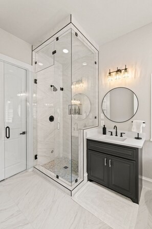 A modern bathroom with a glass-enclosed shower, a round mirror, a white vanity, and dark cabinets. - Executive Townhome in Legacy District Plano & Frisco Welcoming FIFA Guests! (Plano)