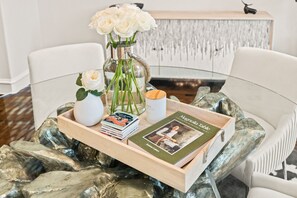 Una mesa de cristal con una bandeja que sostiene un jarrón de rosas blancas, un libro titulado 'Magnolia Table' y una pila de revistas.