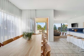 A modern dining area with a long wooden table, chairs, a potted plant, and a kitchen area with stools and a TV. - Ultimate Ocean Views - Pet Friendly sleeps 20 (Jan Juc)