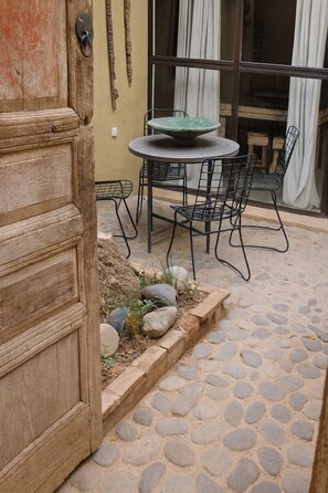 Eine terrasse mit Steinpflaster, ein runder Tisch und Metallstühle, eine Holztür und ein Fenster mit Vorhängen.