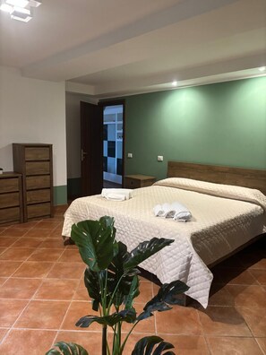 A bedroom with a bed, a wooden dresser, a plant, and a door leading to a bathroom. - Vrbo Property (Castel Gandolfo)