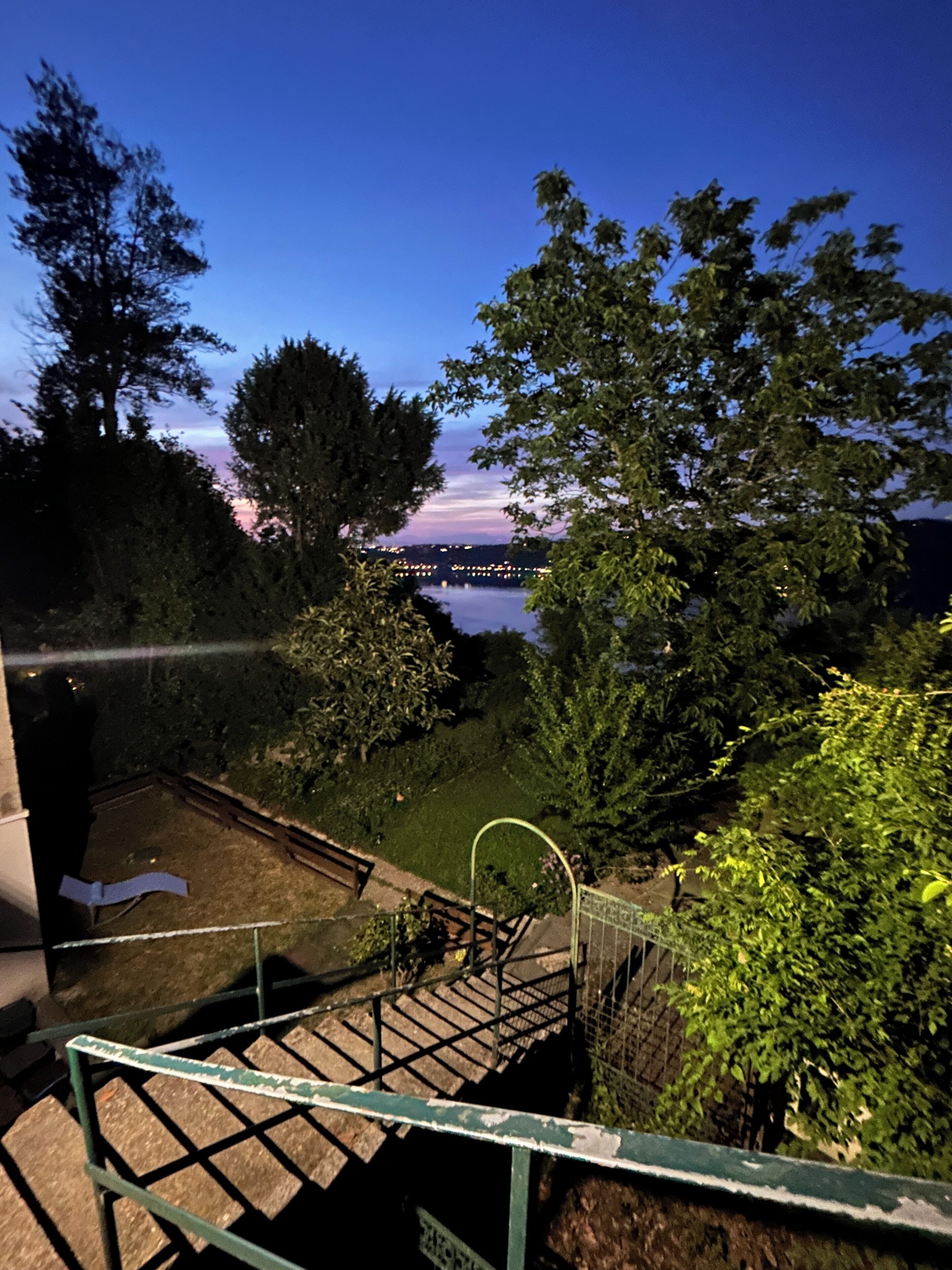 Een trap die leidt naar een gazon met bomen en een uitzicht op een verre stadslijn in de schemering.