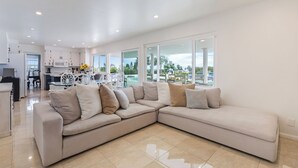 A spacious living room with a large beige sectional sofa, a kitchen in the background, and large windows offering a view of the outdoors. - Skyline Palms by Avantstay Pool + 2 Hot Tubs + Outdoor Dining Near La Jolla + Pacific Beach (San Diego)