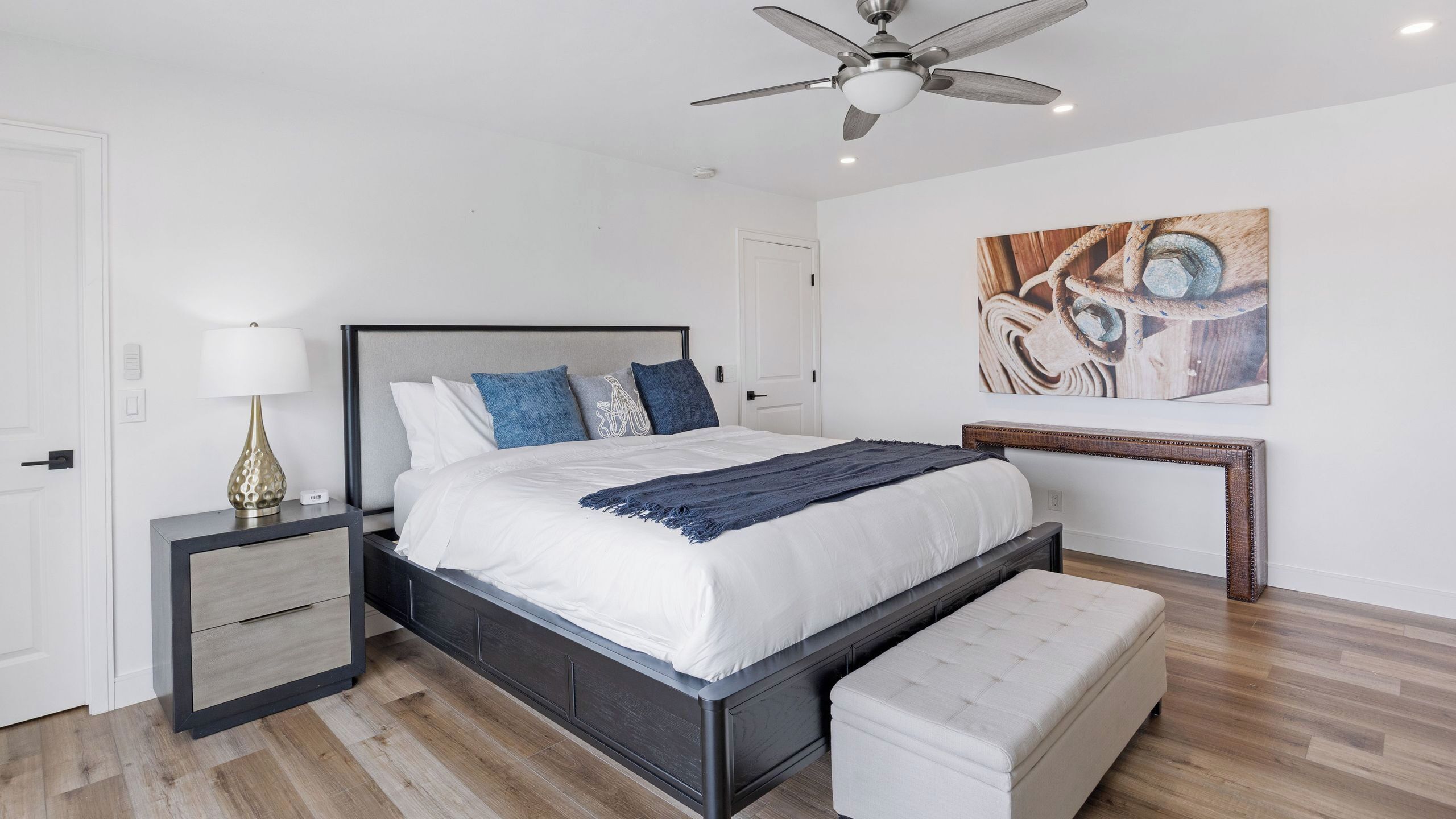 Un dormitorio moderno con cama, mesitas de noche, un banco, ventilador de techo y un cuadro en la pared.