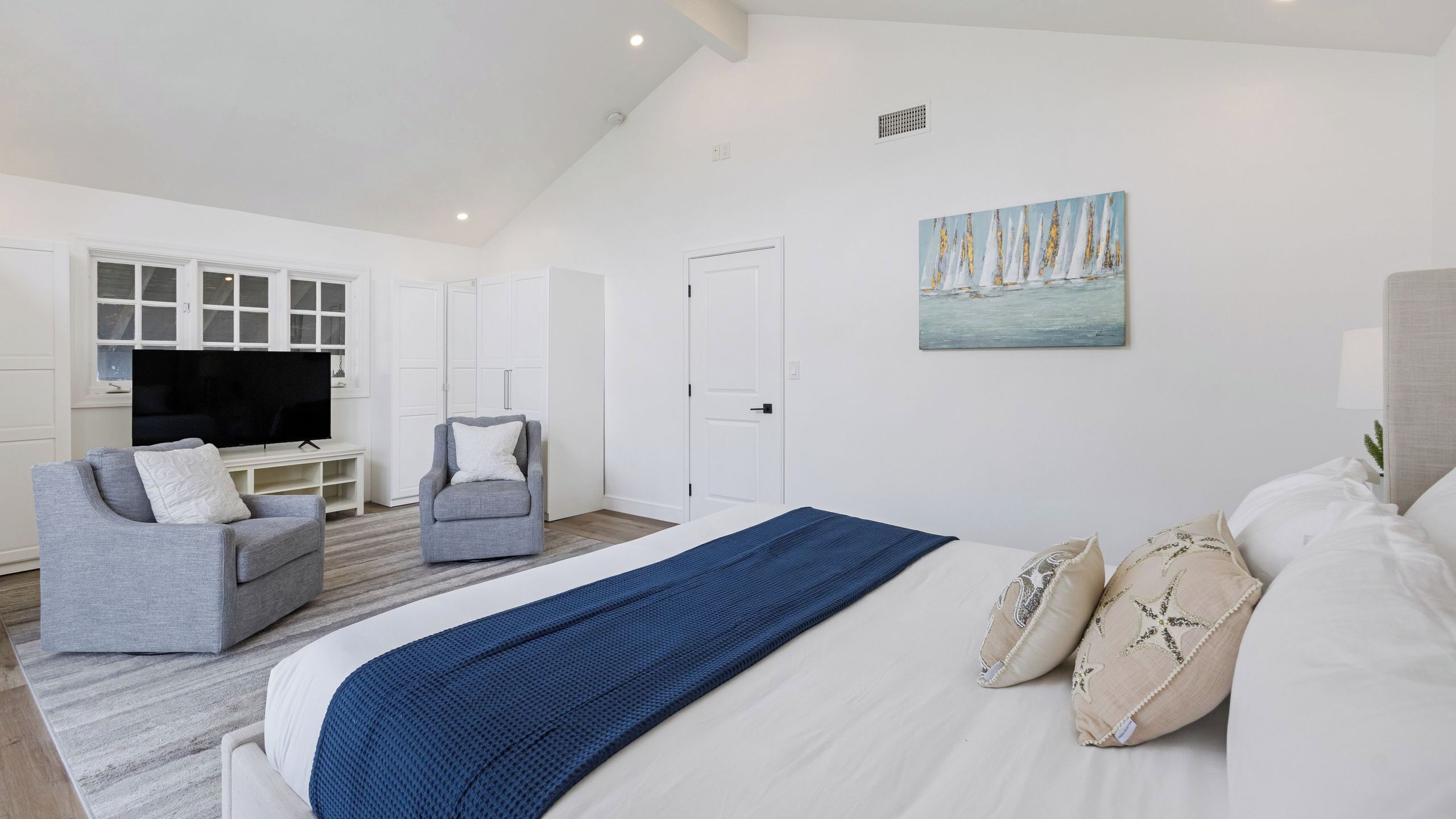 Een moderne slaapkamer met een bed, een grijze fauteuil, een televisie en een schilderij aan de muur.