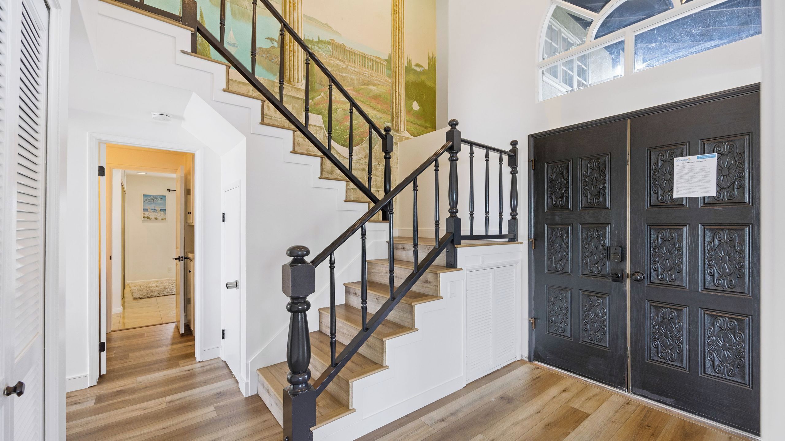 A staircase with black balusters leading to a landing, a dark wooden door with decorative panels, and a mural on the adjacent wall.
