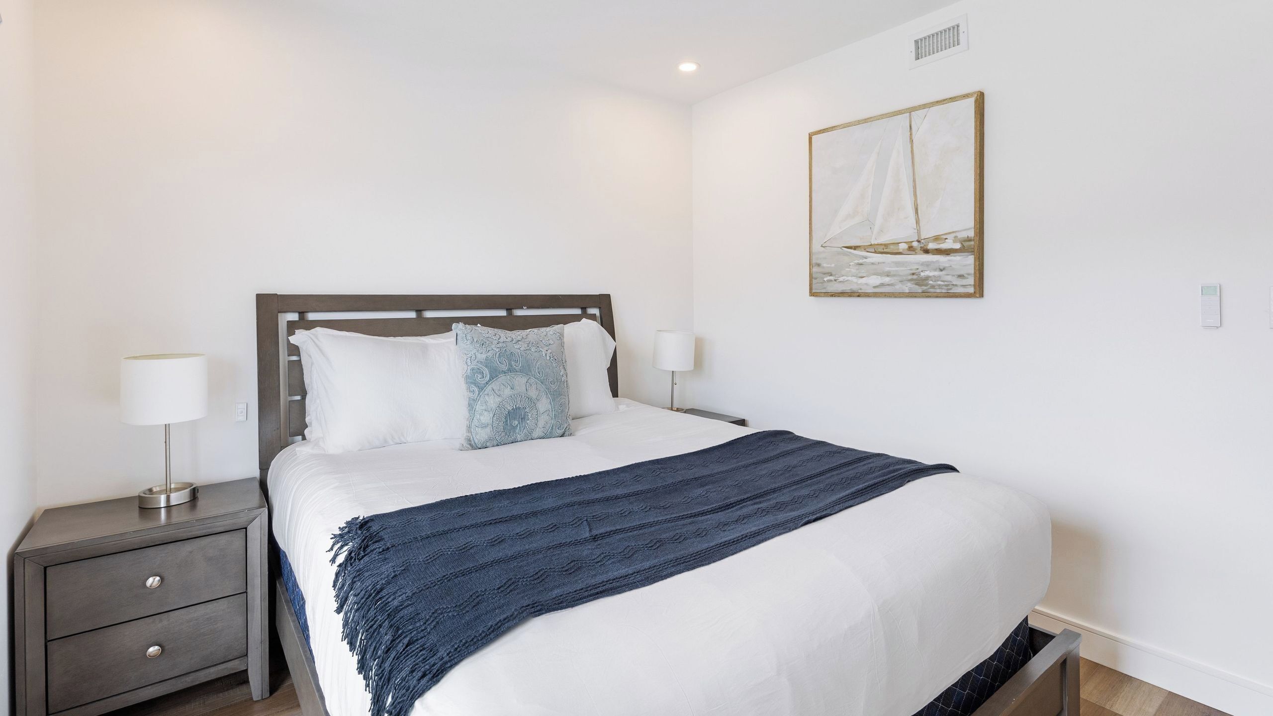 A neatly made bed with a blue blanket, a bedside table with a lamp, and a wall-mounted painting.