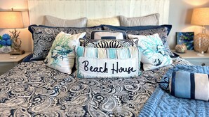 En säng med ett mönstrat överkast, flera kuddar och en dekorativ kudde med texten ”Beach House”.