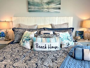 A bed with a patterned bedspread, multiple pillows, and a decorative pillow with 'Beach House' text. - 2BR Holden Beach condo with pool and pet-friendly. (Holden Beach)