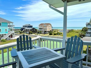 A balcony with Adirondack chairs, a table, and a view of the ocean and beach houses. - 2BR Holden Beach condo with pool and pet-friendly. (Holden Beach)