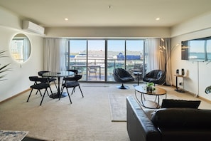 A modern living room with a sofa, a round table, and chairs, a balcony with a view of the city, and a flat-screen TV mounted on the wall. - Harbour Views Morning Sun on Iconic Princes Wharf (Auckland City)