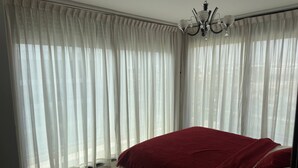 Ein Schlafzimmer mit Bett, weißen Vorhängen, einer Deckenleuchte und Blick durch das Fenster.