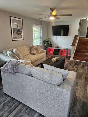 A living room with a grey sofa, a coffee table, a fireplace, a ceiling fan, and a TV mounted on the wall. - River City Retreat •Hot Tub •Game Room• Sleeps 8 Near Fort Benning (Columbus)